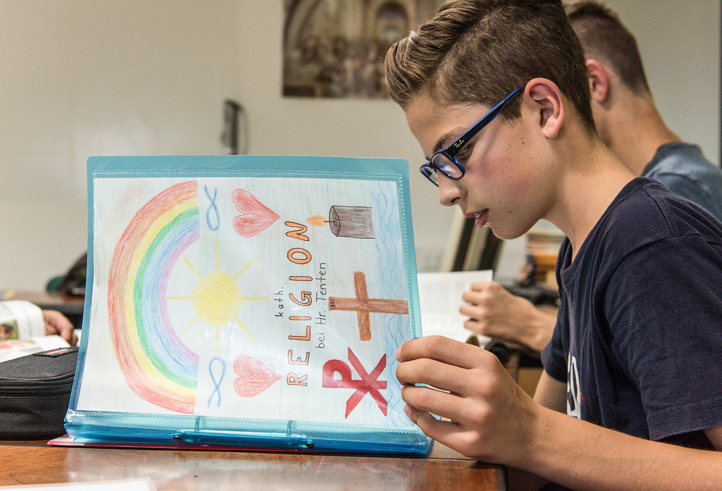 Ein Schüler blättert in seiner Arbeitsmappe beim katholischen Religionsunterricht in einer 8. Klasse. Auf dem Deckblatt sind christliche Symbole gezeichnet - ein Christusmonogramm, ein Kreuz, eine Kerze, Fischsymbole sowie Regenbogen, Sonne und Herzen. (Foto: KNA)